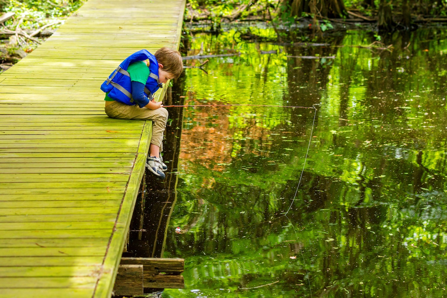 An impatient young, fishing boy stares at his still bobber, willing it to move, as he leans over the edge of the dock he is fishing from at Lake Bistineau, Louisiana.  He is wearing a life jacket.